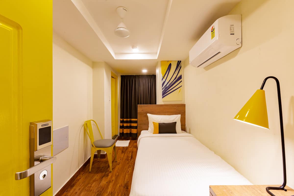 A hotel room with a bed, a desk with a chair, an air conditioning unit, and a wall-mounted lamp.