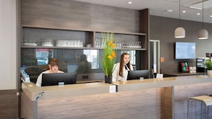 A reception desk with two staff members, a potted plant with orange flowers, and a digital screen displaying information. - Campanile Duisburg City (Duisburg)