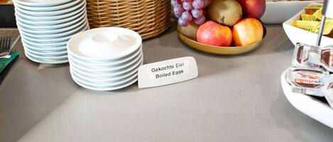 Una mesa de buffet con platos, una cesta de mimbre, un cuenco de frutas y un letrero que indica huevos cocidos.