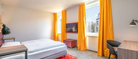 A modern hotel room with a bed, a desk, a chair, and a window with curtains.