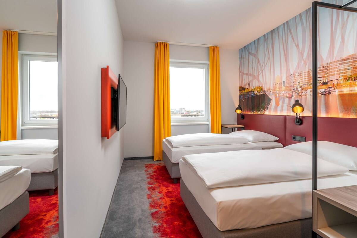 Habitación de hotel con dos camas, un ventanal con cortinas amarillas, vista panorámica de la ciudad y un cuadro en la pared.