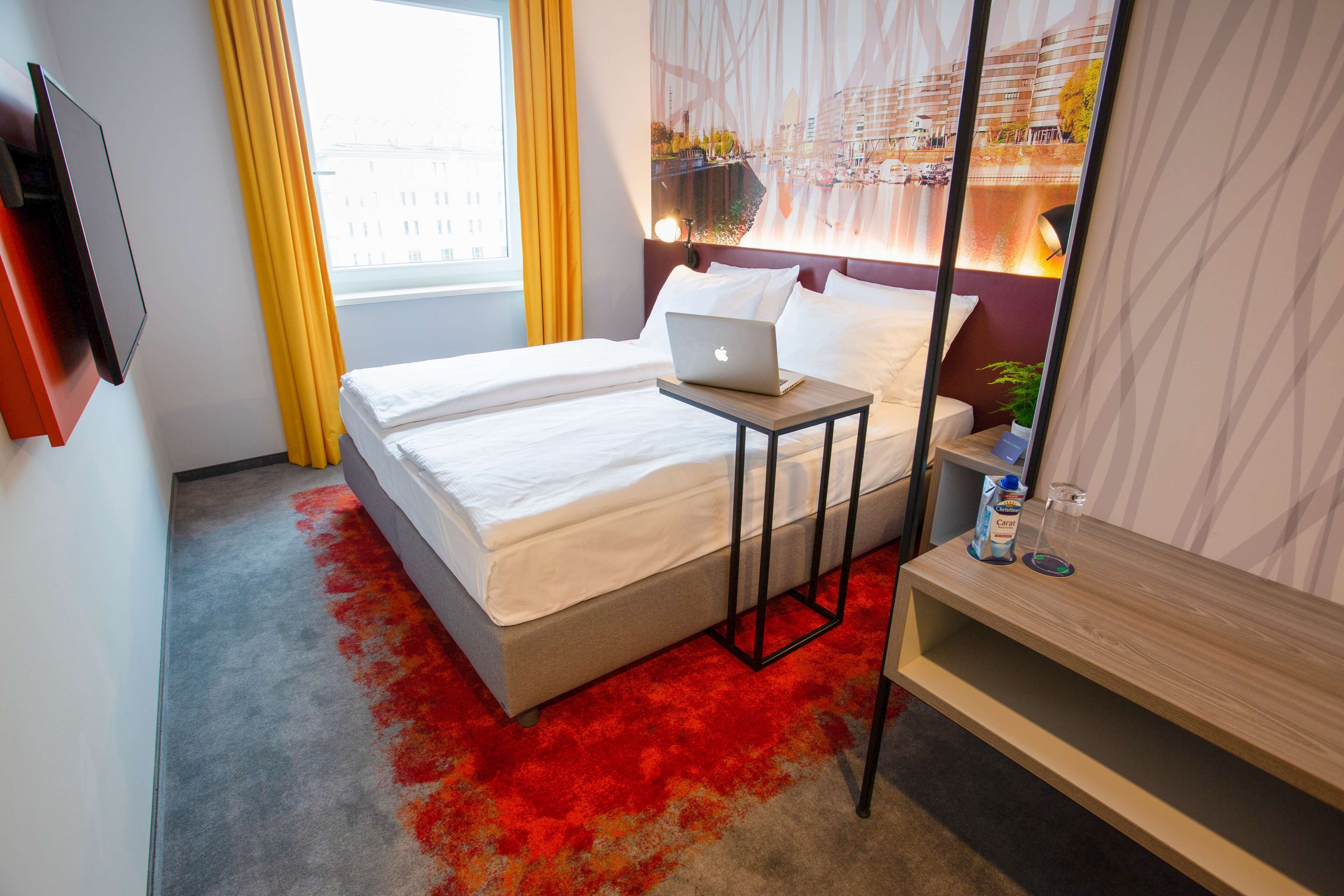 A hotel room with a bed, a laptop on a bedside table, a bedside lamp, a small table with a bottle and glass, and a view of a cityscape through a window with yellow curtains.