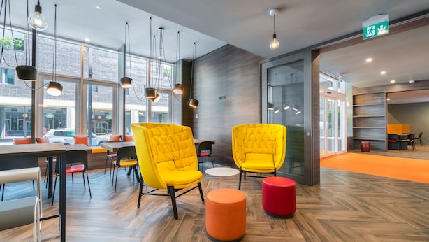 A modern interior with wooden flooring, yellow chairs, and a red ottoman. - Campanile Duisburg City (Duisburg)