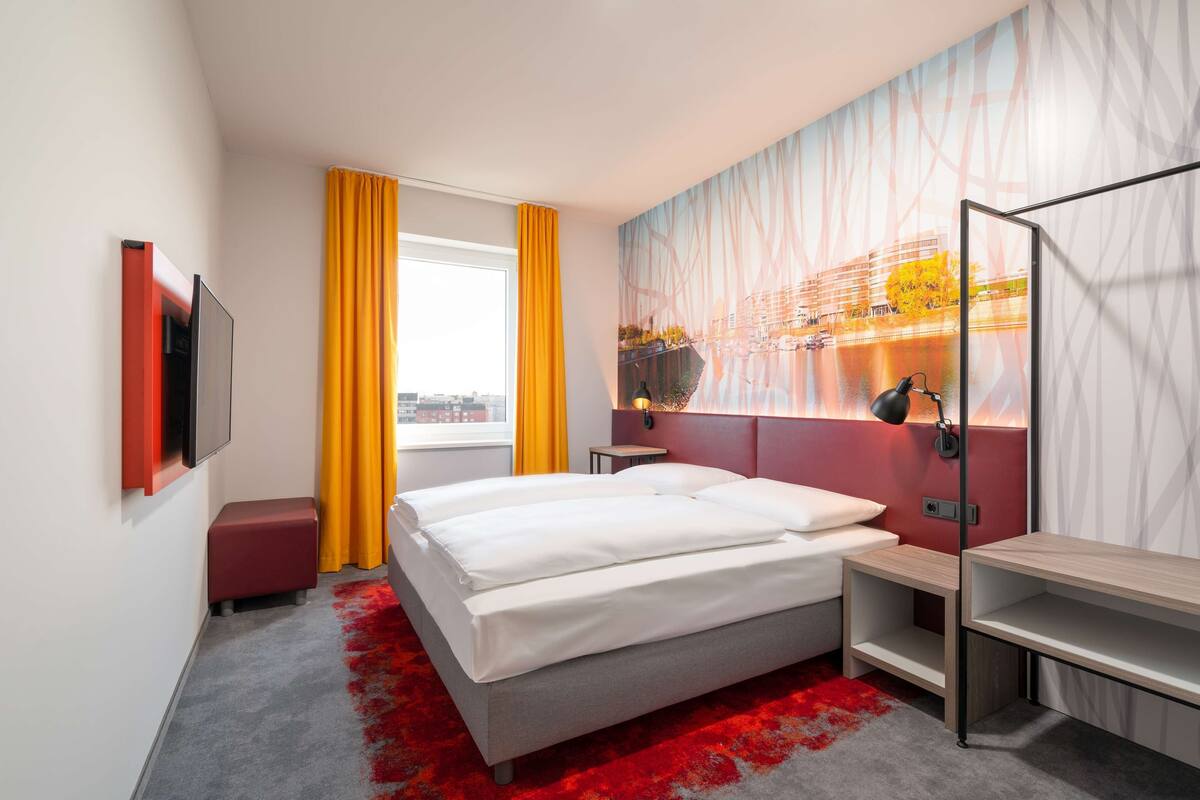 Una habitación de hotel moderna con una cama grande, un escritorio, un televisor y un mural con vista a la ciudad.