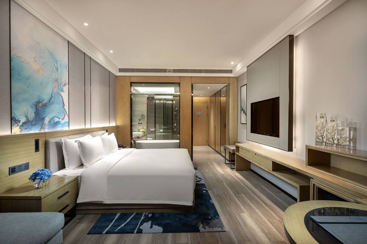 A modern hotel room with a large bed, a TV, a desk, and a bathroom visible through a glass door.