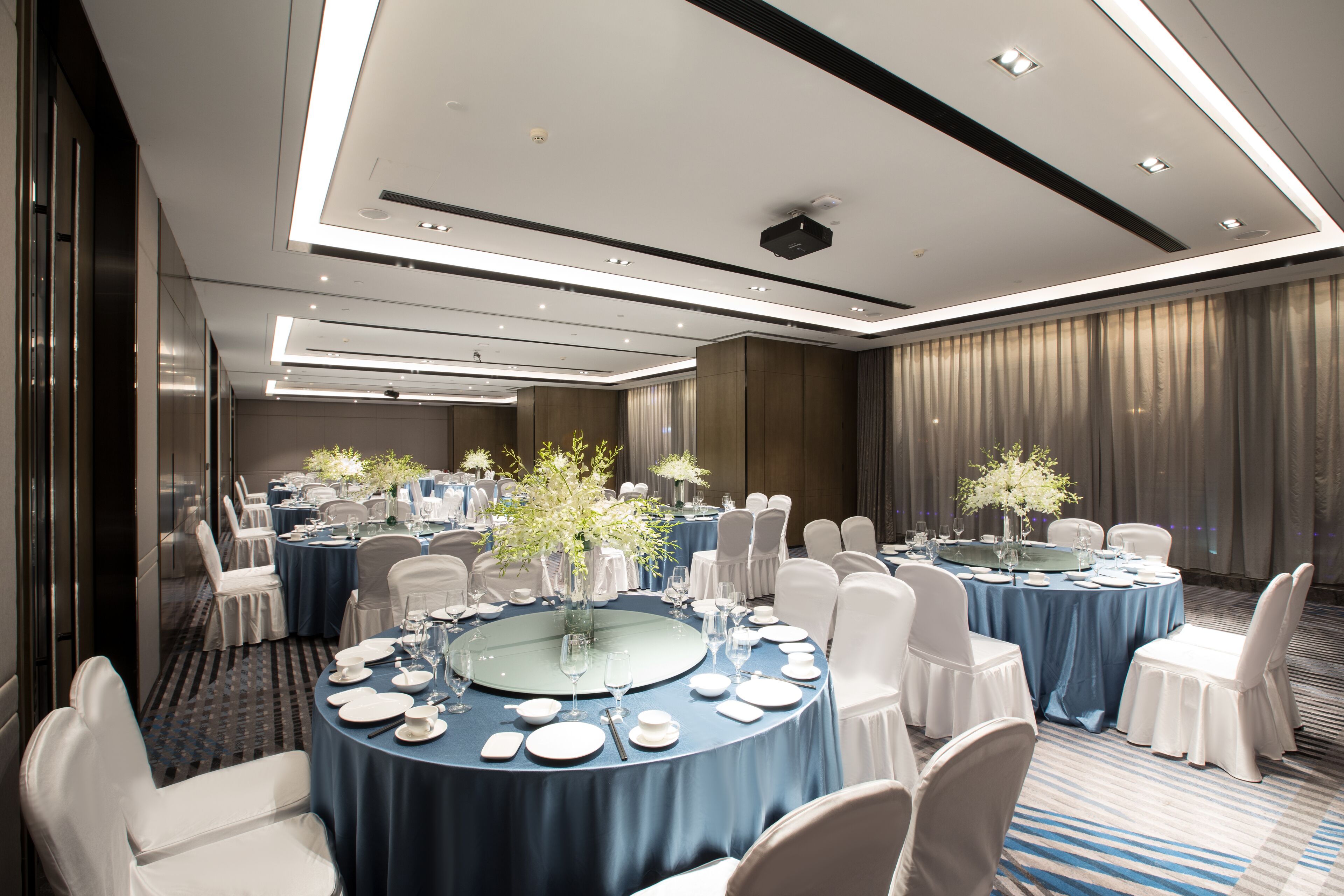 Una sala da banchetti con tavoli rotondi apparecchiati per la cena, sedie bianche e tovaglie blu.