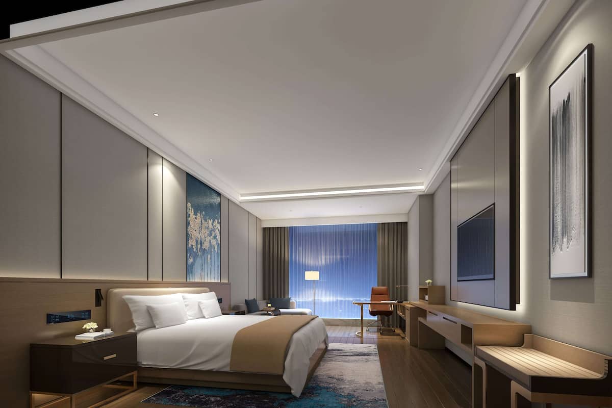 A modern hotel room with a large bed, bedside table, desk, and chair.