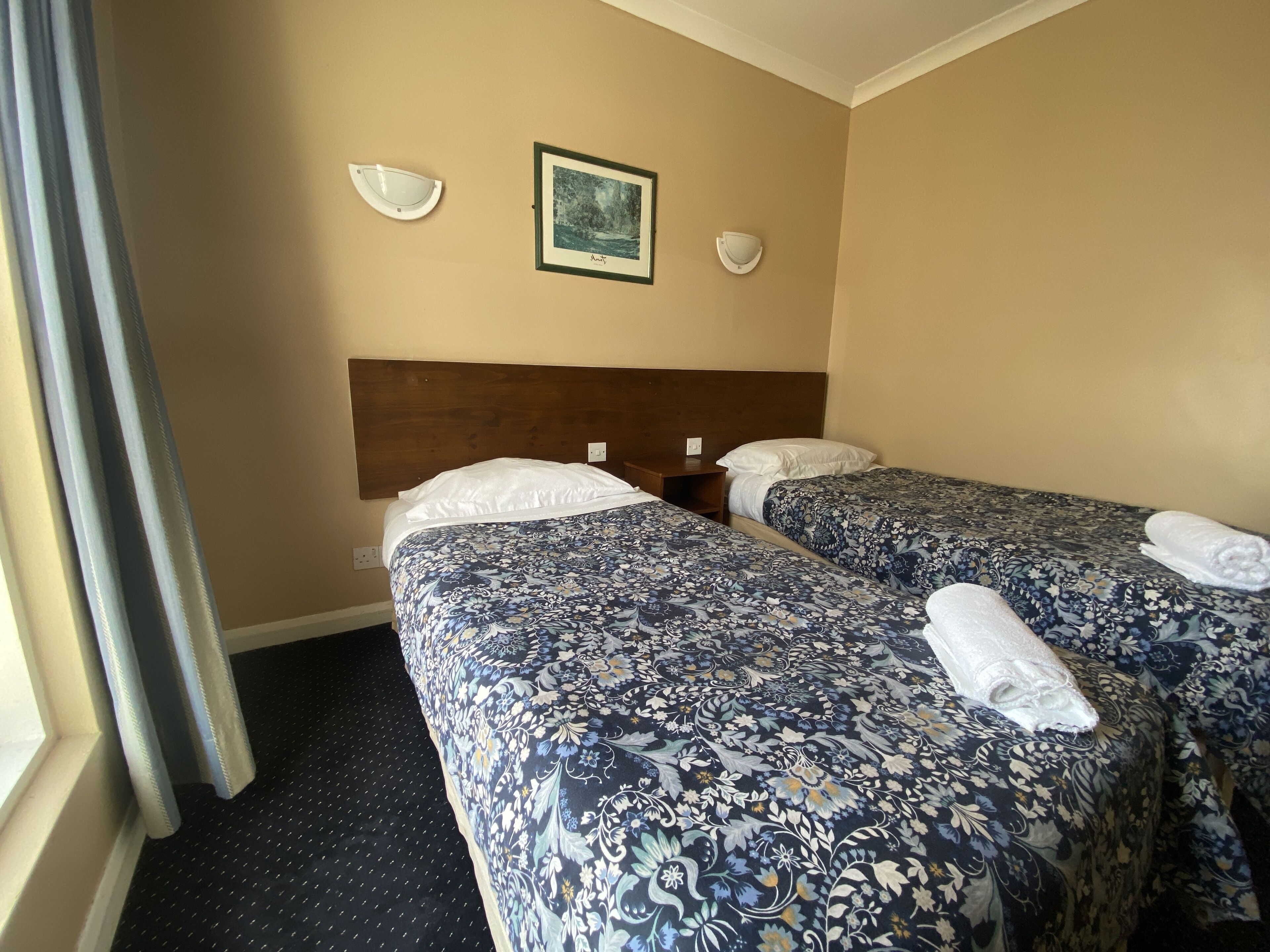 A hotel room with two beds, a wooden headboard, a framed picture on the wall, and a window with curtains.