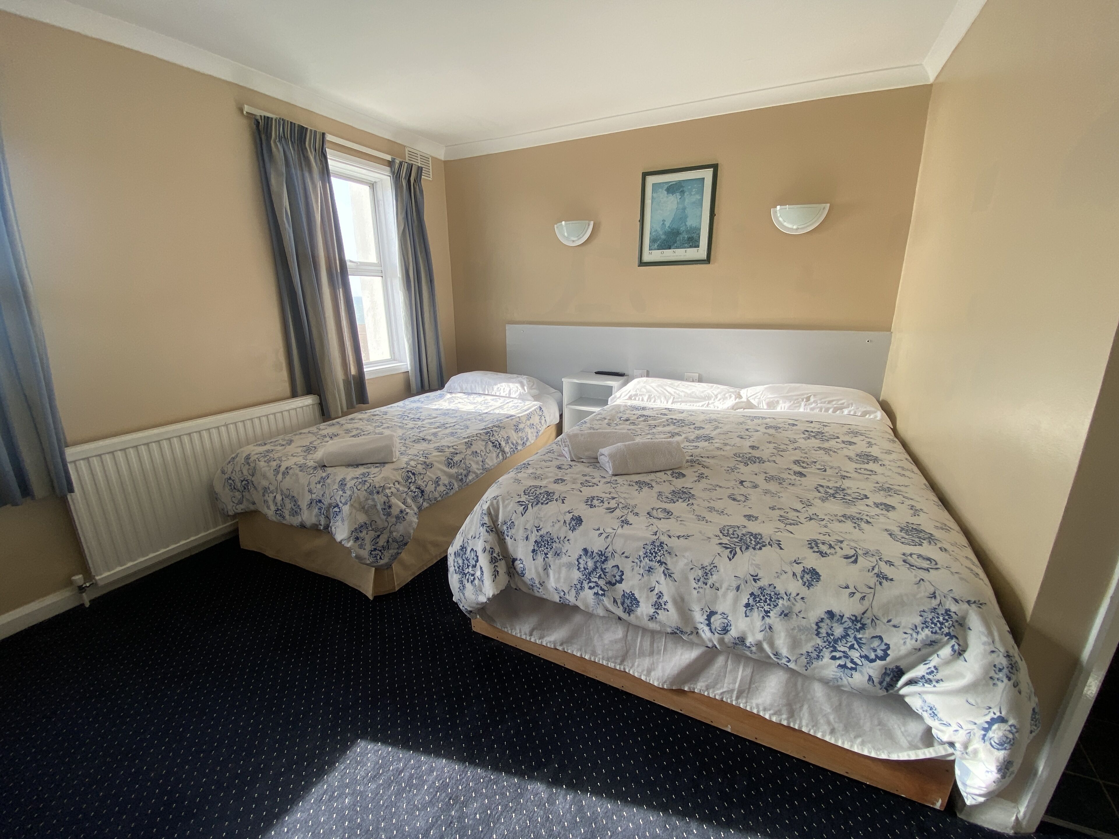 A hotel room with two beds, a radiator, a window with curtains, a framed picture on the wall, and a dark carpet.