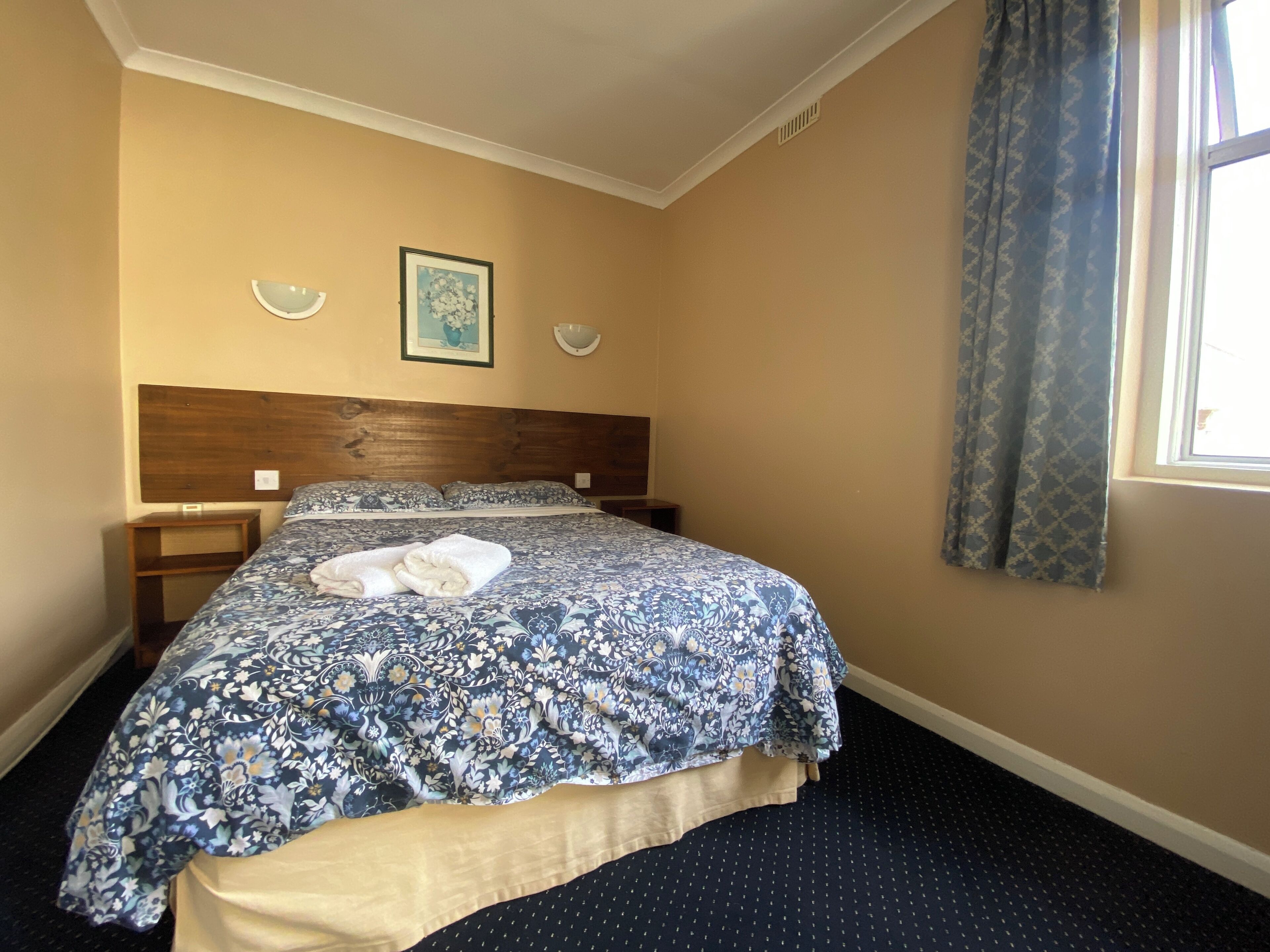 A hotel room with a bed, two bedside tables, a headboard, a window with curtains, and a wall-mounted light fixture.