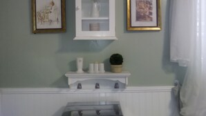 A bathroom with a white cabinet above a shelf holding toiletries, a framed picture on the wall, and a potted plant.