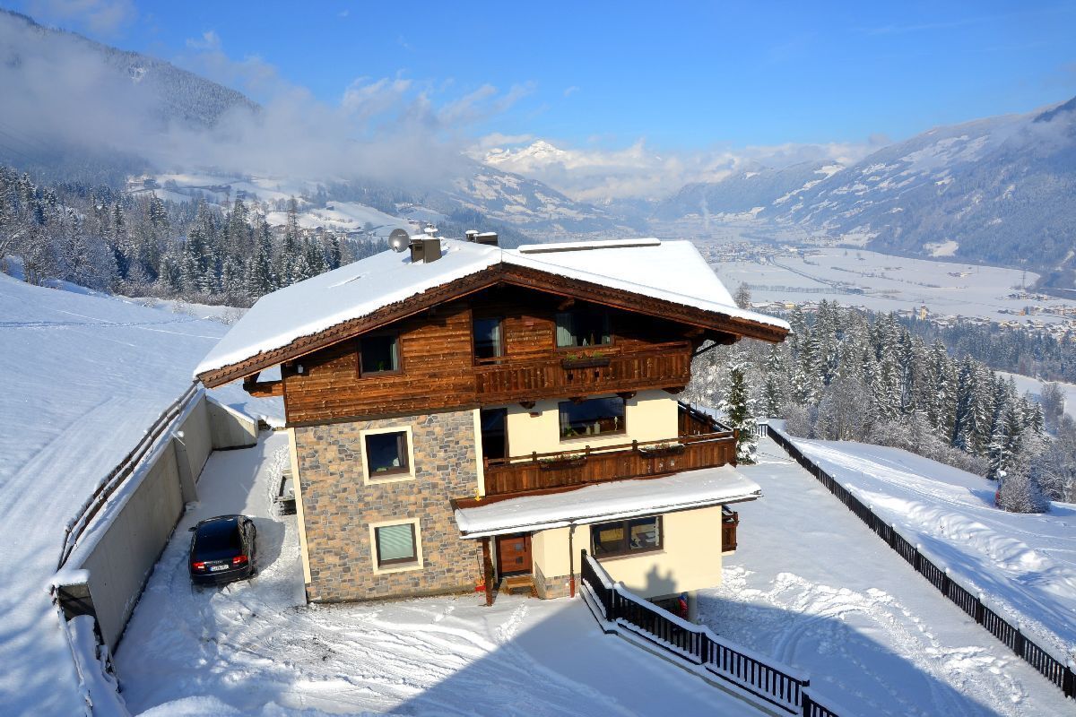 A two-story chalet with a stone facade and wooden upper levels, set against a snowy mountain backdrop.