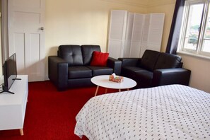 A hotel room with a red carpet, two black leather sofas, a white bed with a patterned cover, a small white table, and a TV on a white stand.