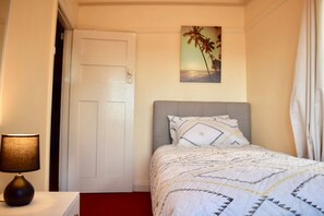 A bedroom with a bed, a nightstand, a lamp, a door, and a wall painting of palm trees.