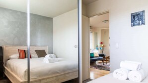A modern hotel room with a large bed, a wooden headboard, and a glass partition. There is a bathroom visible through the partition, and a small table with towels on the right side of the room. - Charming 1 Bedroom Apartment in Vibrant South Yarra (South Yarra)