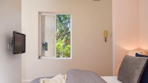 Ein Schlafzimmer mit Bett, einem Fenster mit Blick auf Bäume, einem an der Wand montierten Fernseher und einer Nachttischlampe.