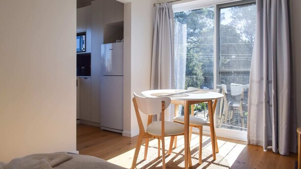 Ein Zimmer mit Esstisch und Stühlen, eine Küche im Hintergrund und ein großes Fenster mit Vorhängen.