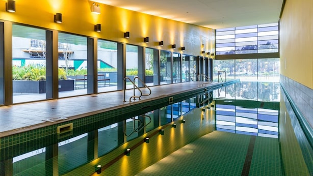 A long indoor swimming pool with clear glass walls, a tiled floor, and overhead lighting. - Modern 1 Bedroom in Tranquil Parklands (Parkville)