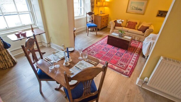 Un coin repas avec une table en bois dressée pour quatre personnes, un tapis rouge à motifs et un canapé en arrière-plan.