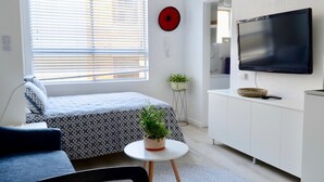 Ein modernes, helles Zimmer mit einem Bett, einem kleinen Tisch mit einer Pflanze, einem Fernseher an der Wand und einem weißen Schrank.
