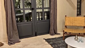 Ein Zimmer mit einer dunklen Holztür, einem gewebten Stuhl, einem runden Tisch mit Gläsern und einem hängenden Kleiderständer.