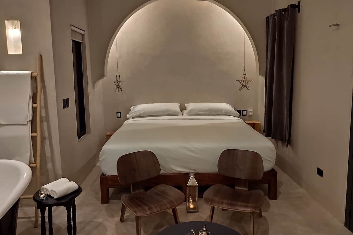 A modern bedroom with a large bed, two wooden chairs, a small table, a ceiling fan, and a nightstand with a lamp.