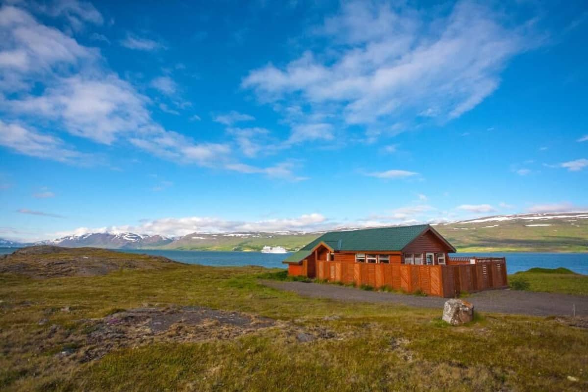 Une cabane en bois avec un toit vert, située sur une colline herbeuse d’où l’on a vue sur un lac et des montagnes.