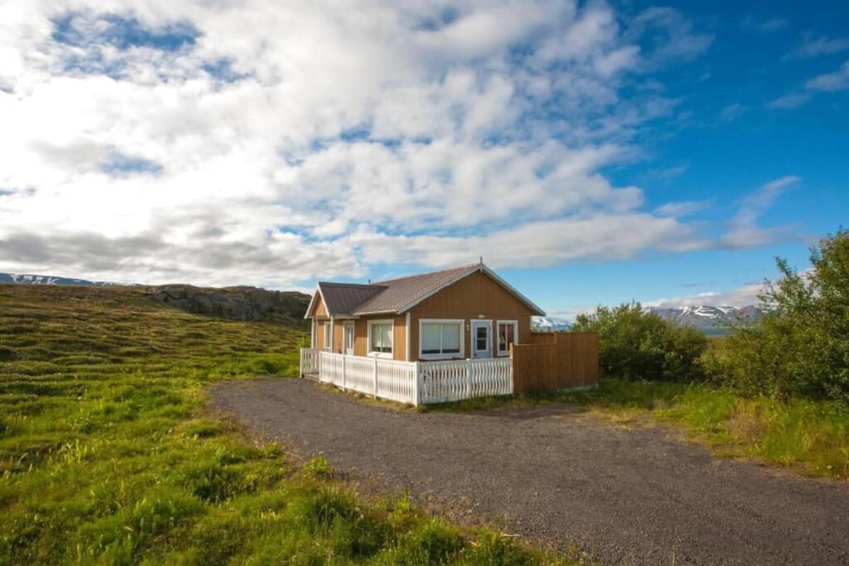 Une maison de plain-pied avec une clôture en bois peinte en blanc, une allée en gravier et un champ verdoyant au premier plan.