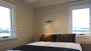A bedroom with a bed, two bedside tables, a nightstand, and a window with a view of the sea. - Vacation Home Fagravik (Akureyri)
