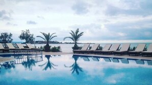 Une piscine bordée de chaises longues et de palmiers, dont l’eau reflète le ciel.