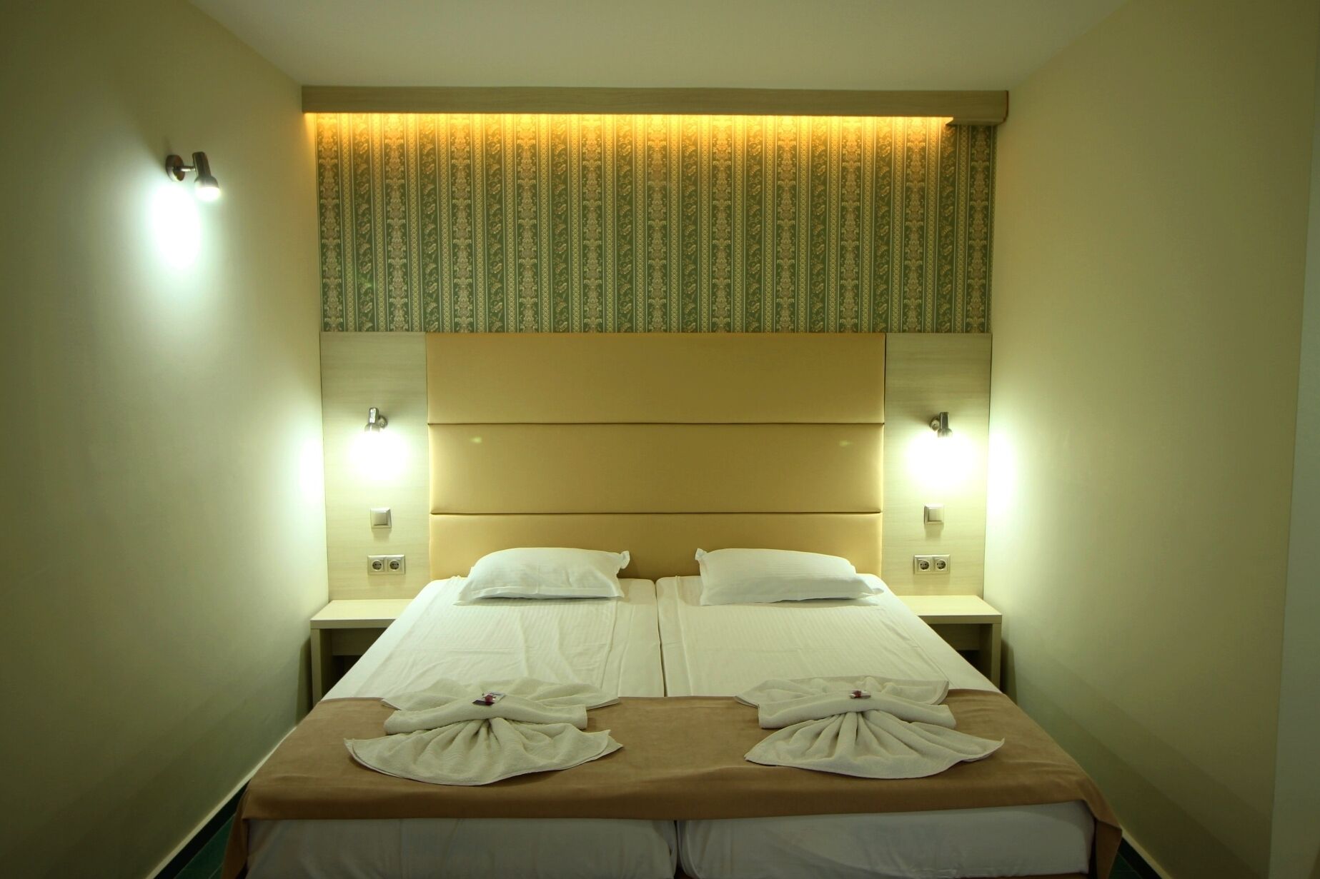Habitación de hotel con dos camas, cabeceros con cortina estampada, lámparas de noche y toallas sobre las camas.