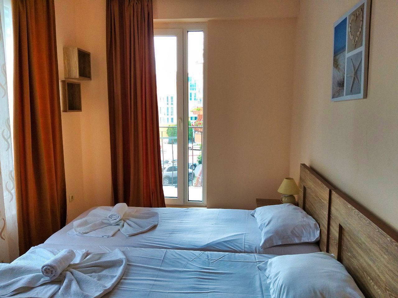 A hotel room with two beds, a window with curtains, a wall clock, and a small table with a lamp.