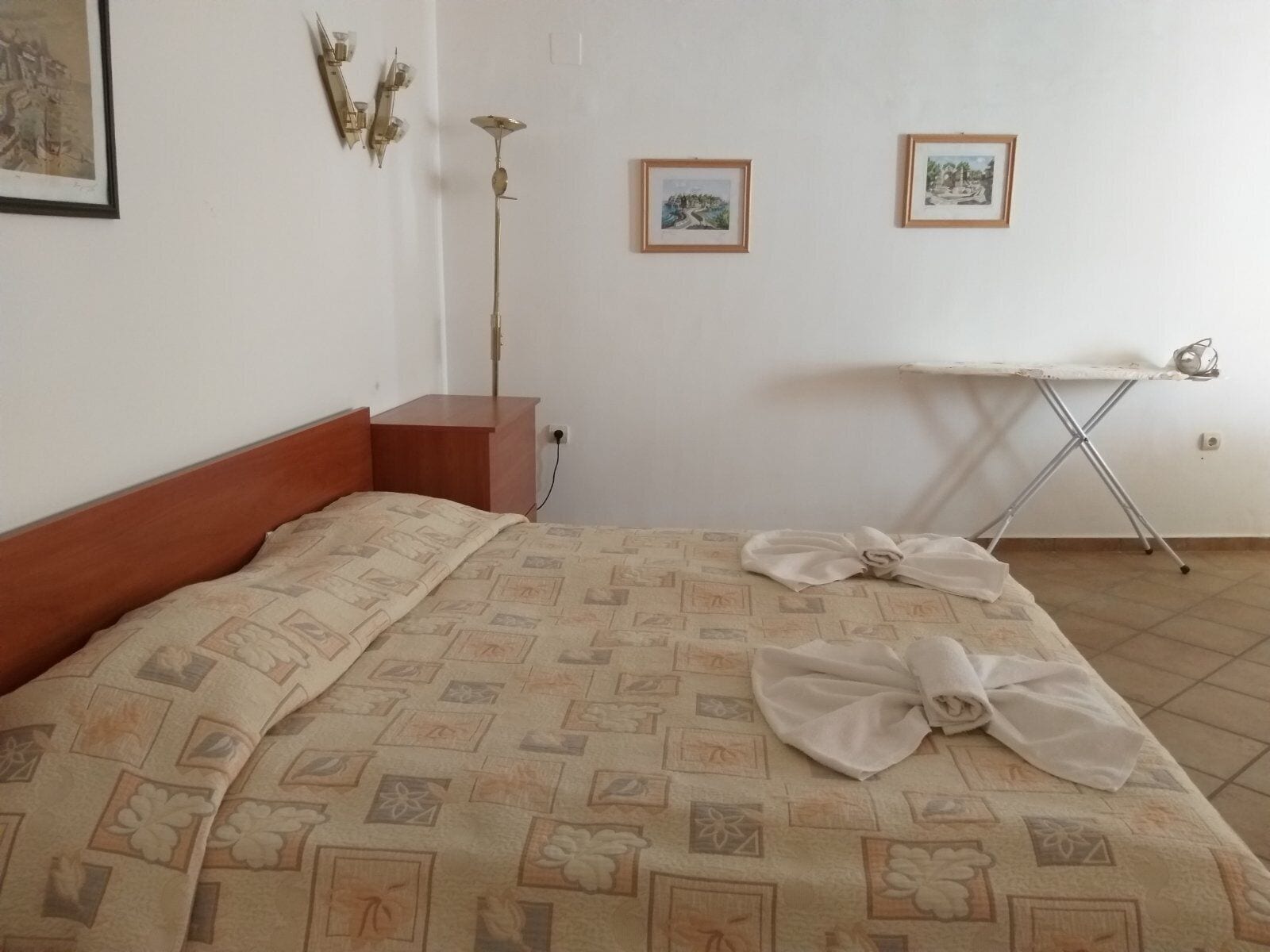 Una cama bien tendida con una colcha estampada, una mesita de noche de madera, una lámpara de pie y dos cuadros enmarcados en la pared.
