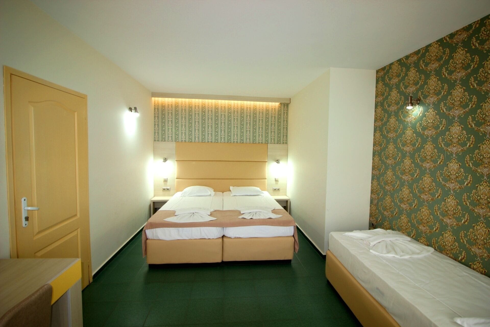 Una habitación de hotel con una cama doble, una cama individual, una puerta de madera y un papel tapiz estampado en una pared.