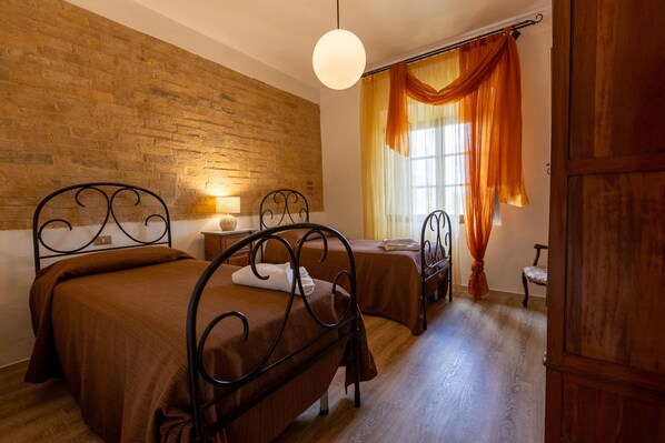 Una habitación con dos camas, una pared de ladrillo, una puerta de madera y una ventana con cortinas naranjas.