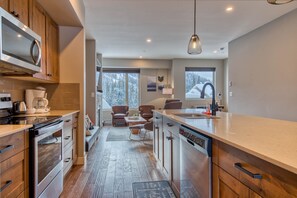 A modern kitchen with wooden cabinets, a dishwasher, and a large island. There is a dining area with a table and chairs, and a view of a snowy landscape through the windows.