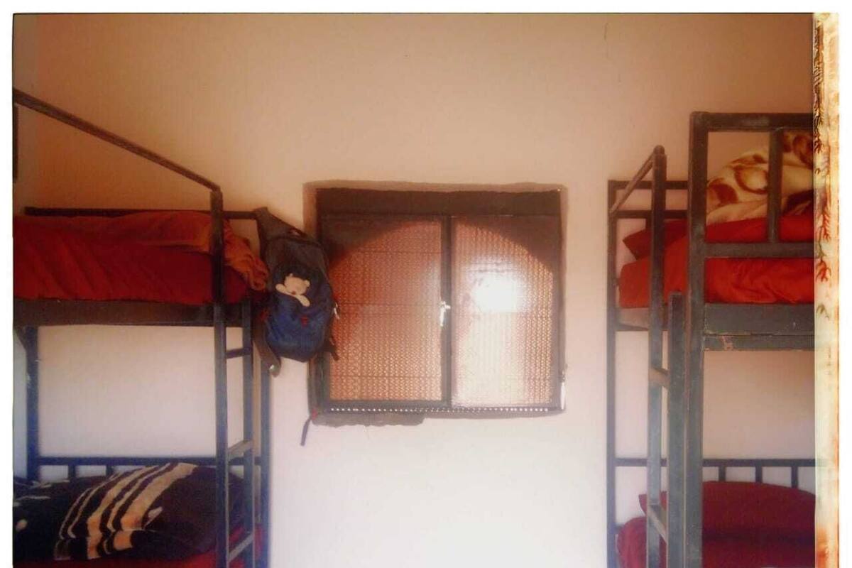 A dormitory room with bunk beds, a window with a mesh screen, and a backpack hanging on the wall.