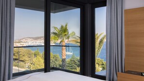 Habitación de hotel con un ventanal grande que ofrece vistas a un pueblo costero, una palmera y un velero en el agua.