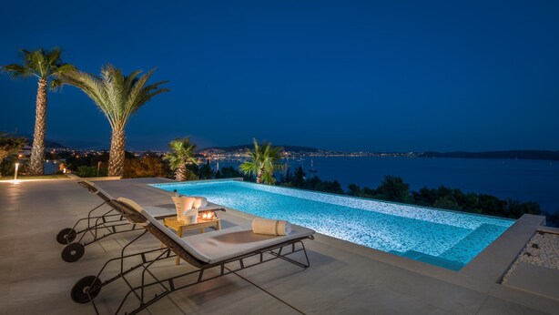 Una piscina infinita con sillones reclinables y una botella de Moët champagne, con vista a un paisaje urbano costero de noche.
