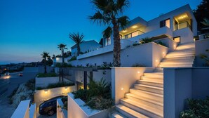 Una casa moderna de varios pisos con una escalera destacada, iluminación exterior y un automóvil en la entrada.