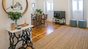 Un vaste espace de vie doté d’un plancher en bois, d’un grand miroir, d’une table à manger avec des chaises, d’un téléviseur et de deux poufs.