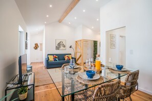 A modern living room with a glass dining table, wicker chairs, a blue sofa, and a television on a glass stand.