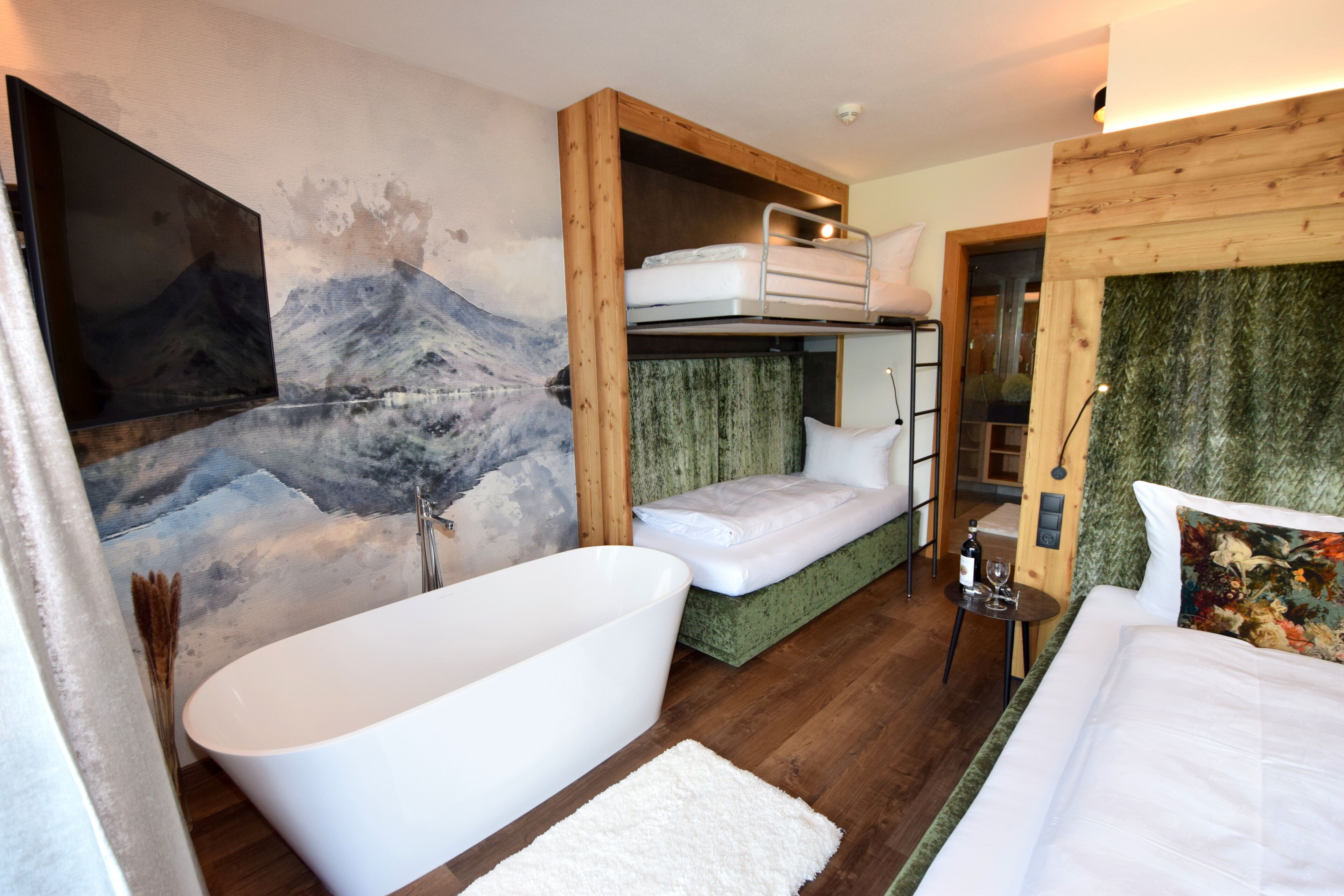 A modern hotel room with a large bathtub, bunk beds, a painting of a mountain, and a green textured wall.