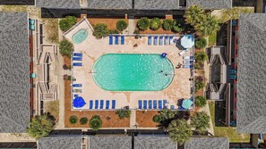 Een luchtbeeld van een resort met een centraal zwembad, meerdere loungegebieden met parasols en omlijstende palmbomen.