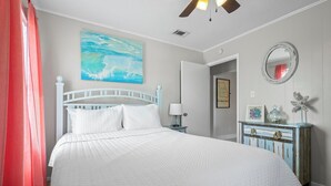 Un dormitorio con cama blanca, ventilador de techo, un cuadro, cómoda y espejo.