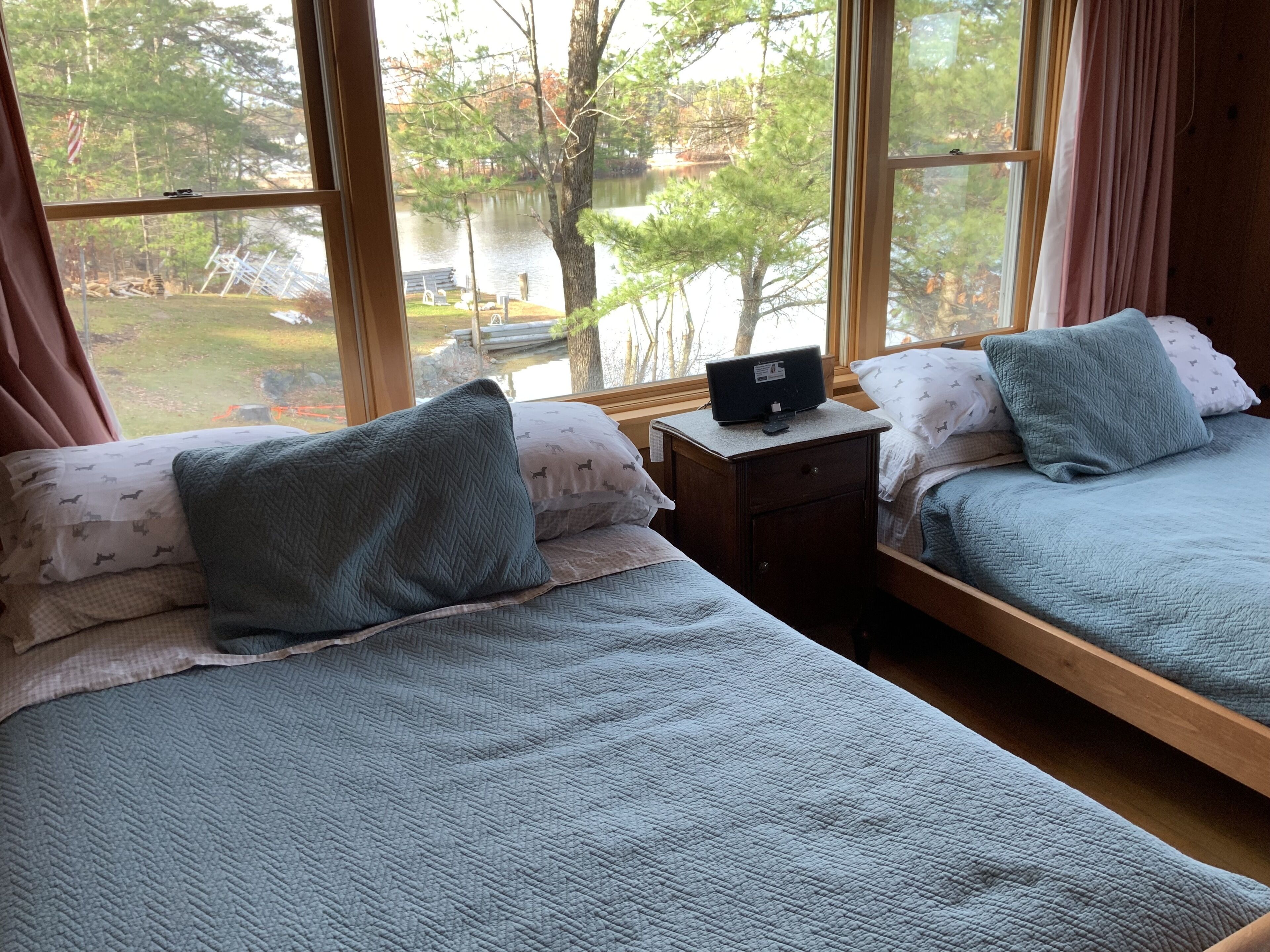 A bedroom with two beds, each with a blue quilted bedspread and matching pillows. There is a small wooden nightstand with a television between the beds. The room has large windows offering a view of a lake and trees.