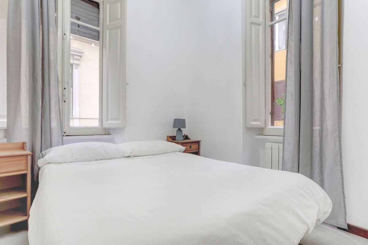 Un dormitorio con cama, mesita de noche, lámpara y ventana con cortinas.
