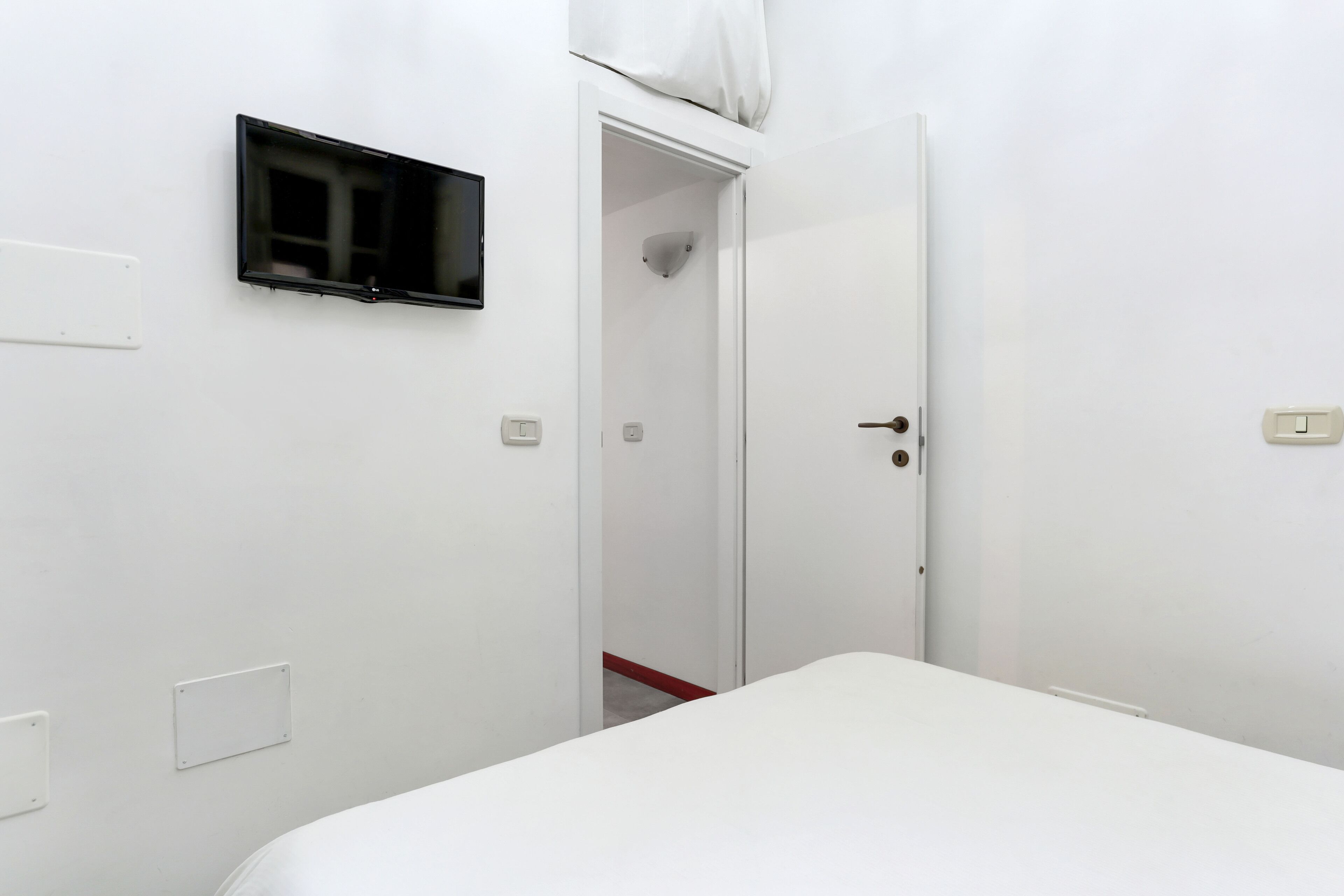 A white room with a bed, a door, and a wall-mounted light fixture.