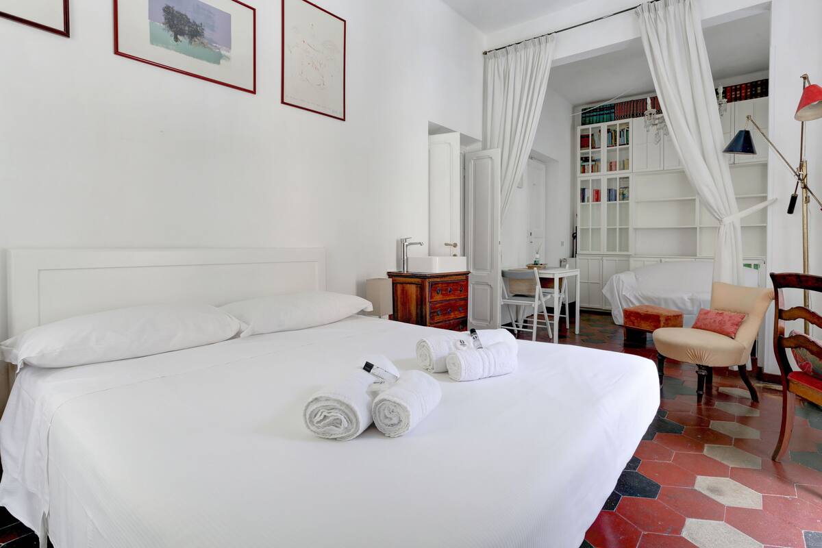 Habitación de hotel con una cama grande, cortinas blancas, una estantería, un escritorio y una silla.