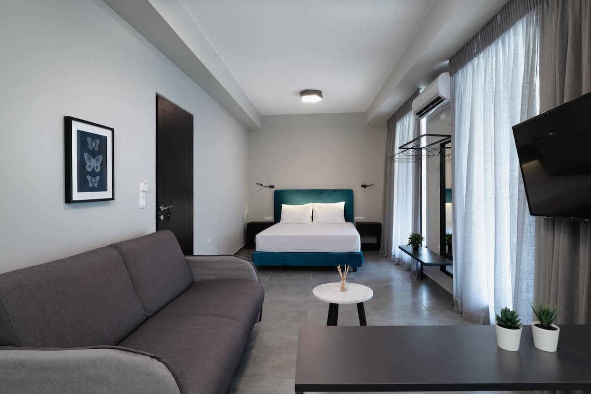 Une chambre d’hôtel moderne avec un canapé gris, un lit avec une literie de couleur turquoise, une petite table ronde et un téléviseur placé sur le mur de droite.